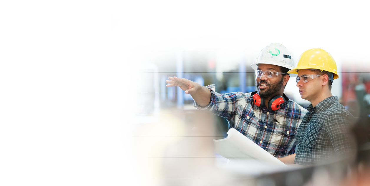 Two multi-ethnic workers in their 30s talking in a metal fabrication plant wearing hardhats and protective eyewear  The man pointing is African-American and his coworker is Hispanic 